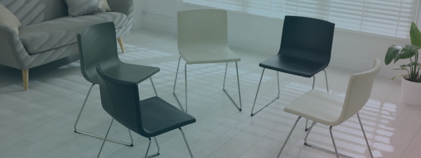 A bright room with 5 chairs arranged in a circle on a pale wooden floor, sunlight filters through blinds with a sofa in the background and a plant to the right.